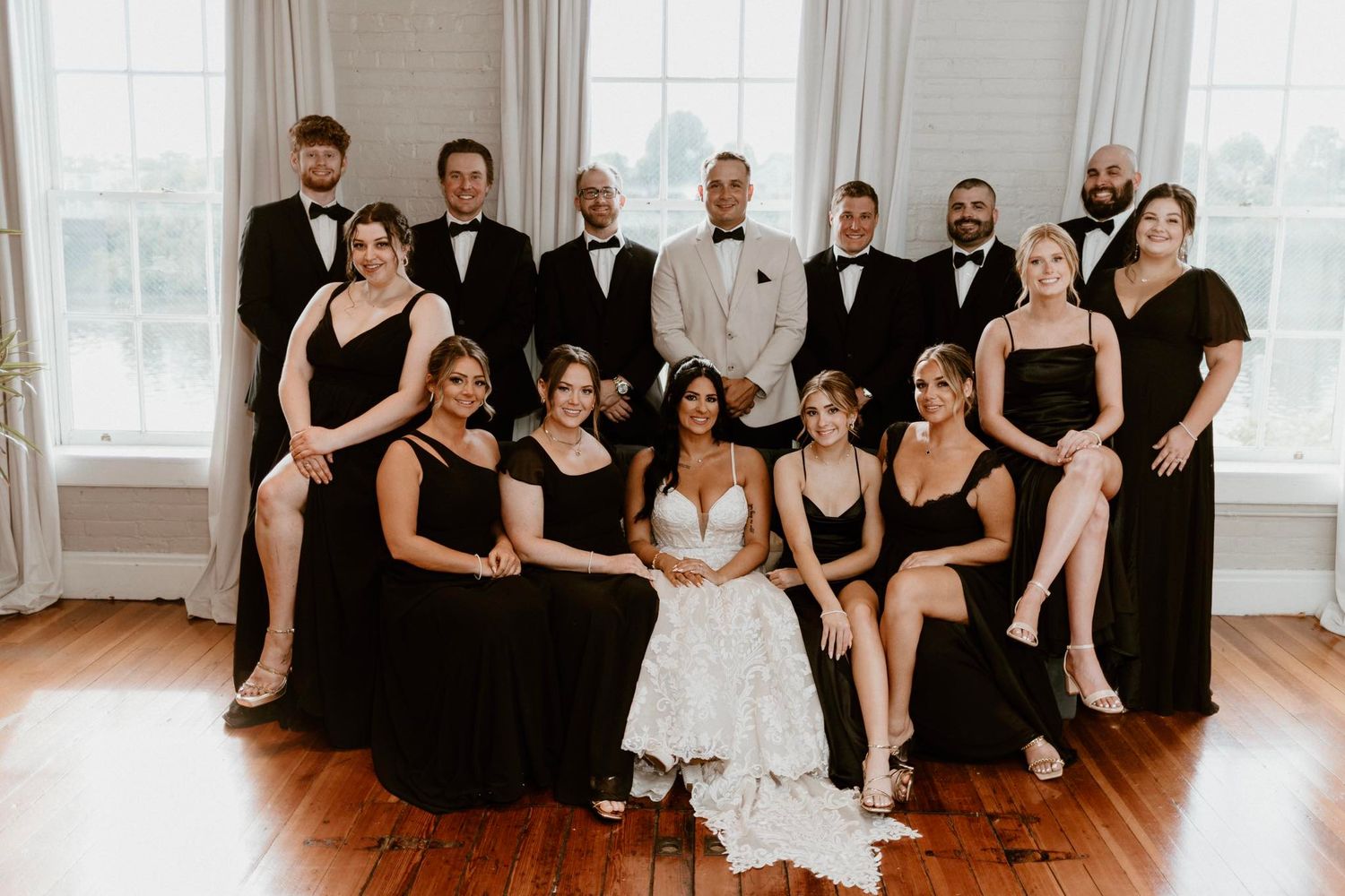 Wedding party in black attire and white wedding dress pose together in elegant room with wooden floors and bright windows.