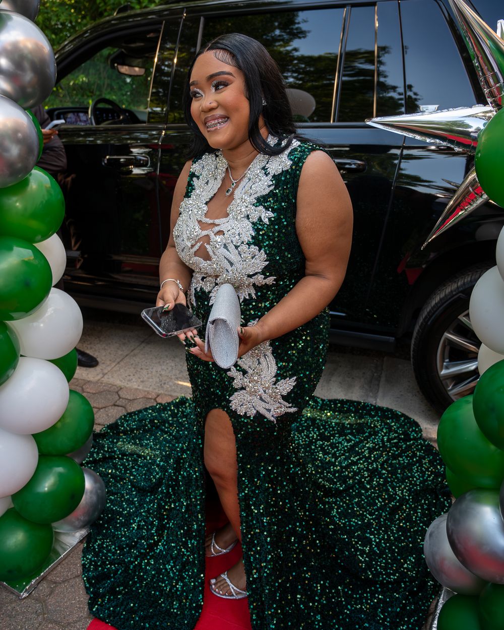 A woman in a sparkly green gown poses by a car with balloon decorations.