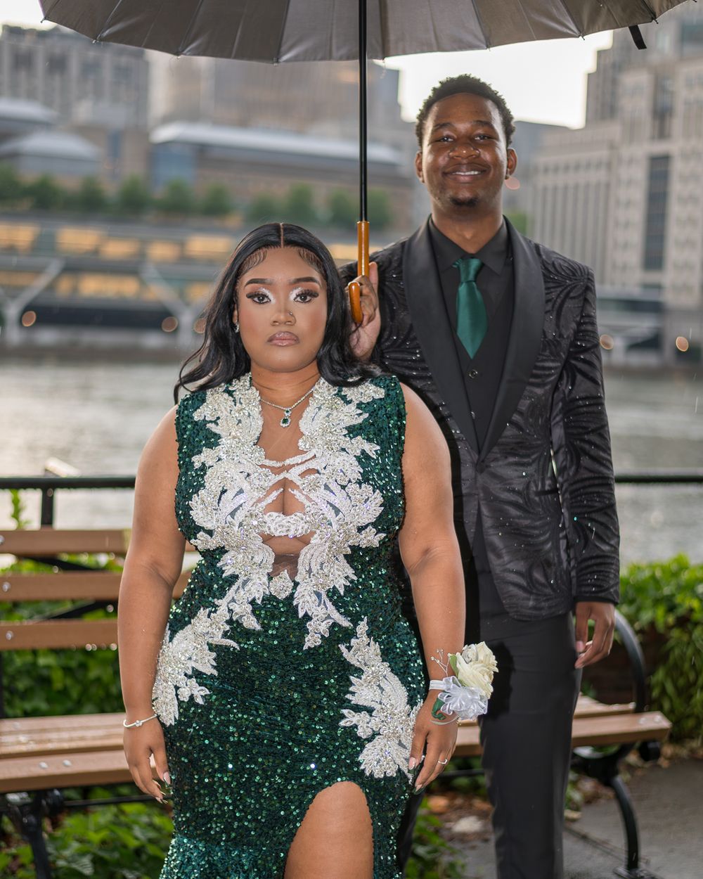 A couple in formal attire poses under an umbrella near a waterfront.