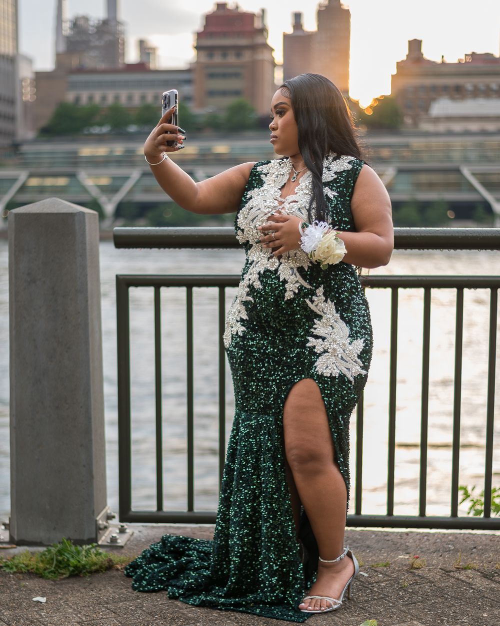 A person in a sequined green gown takes a selfie by a waterfront railing at sunset.