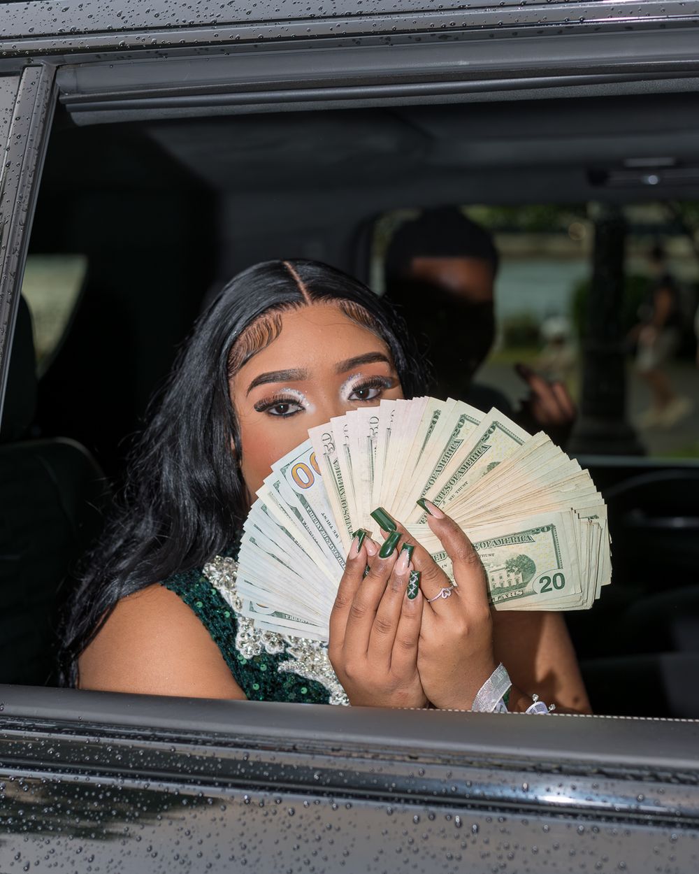 A person in a green dress displays cash while sitting in a car window.