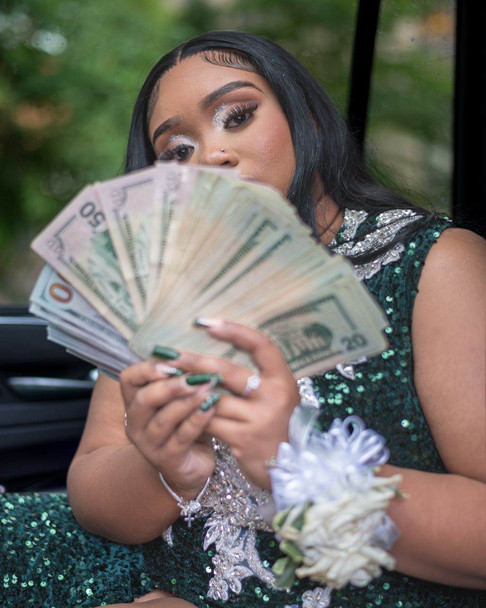 A person in a sparkly dress holds up a fan of dollar bills while seated in a vehicle.