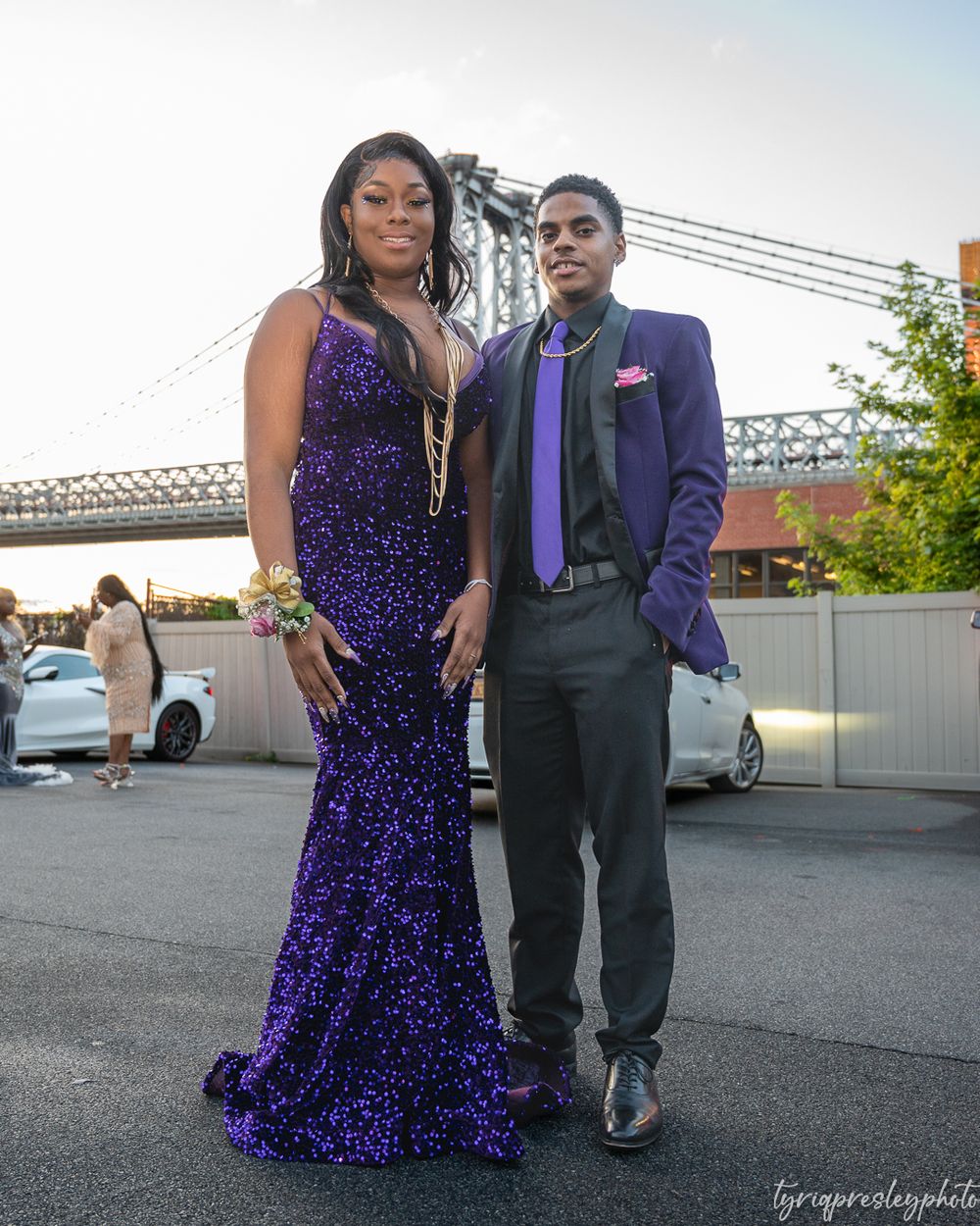 A couple poses in formal purple attire with a bridge visible in the background.