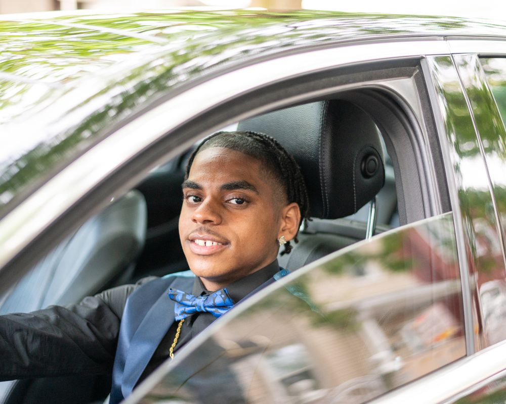 Driver in formal attire with bow tie smiling from inside vehicle.