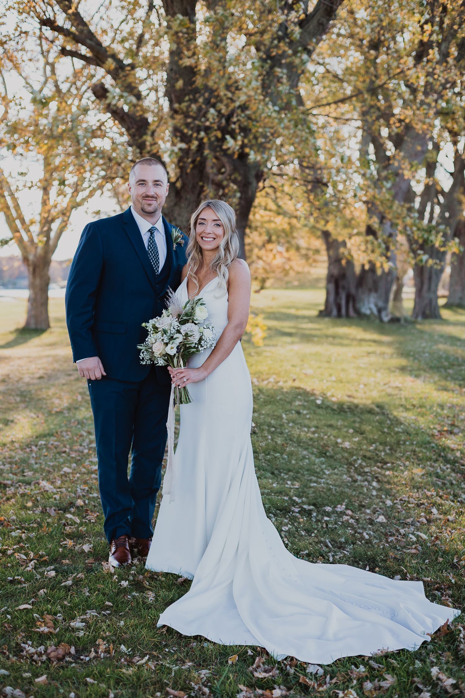 A couple poses outdoors on their wedding day in a fall setting with autumn trees and foliage.