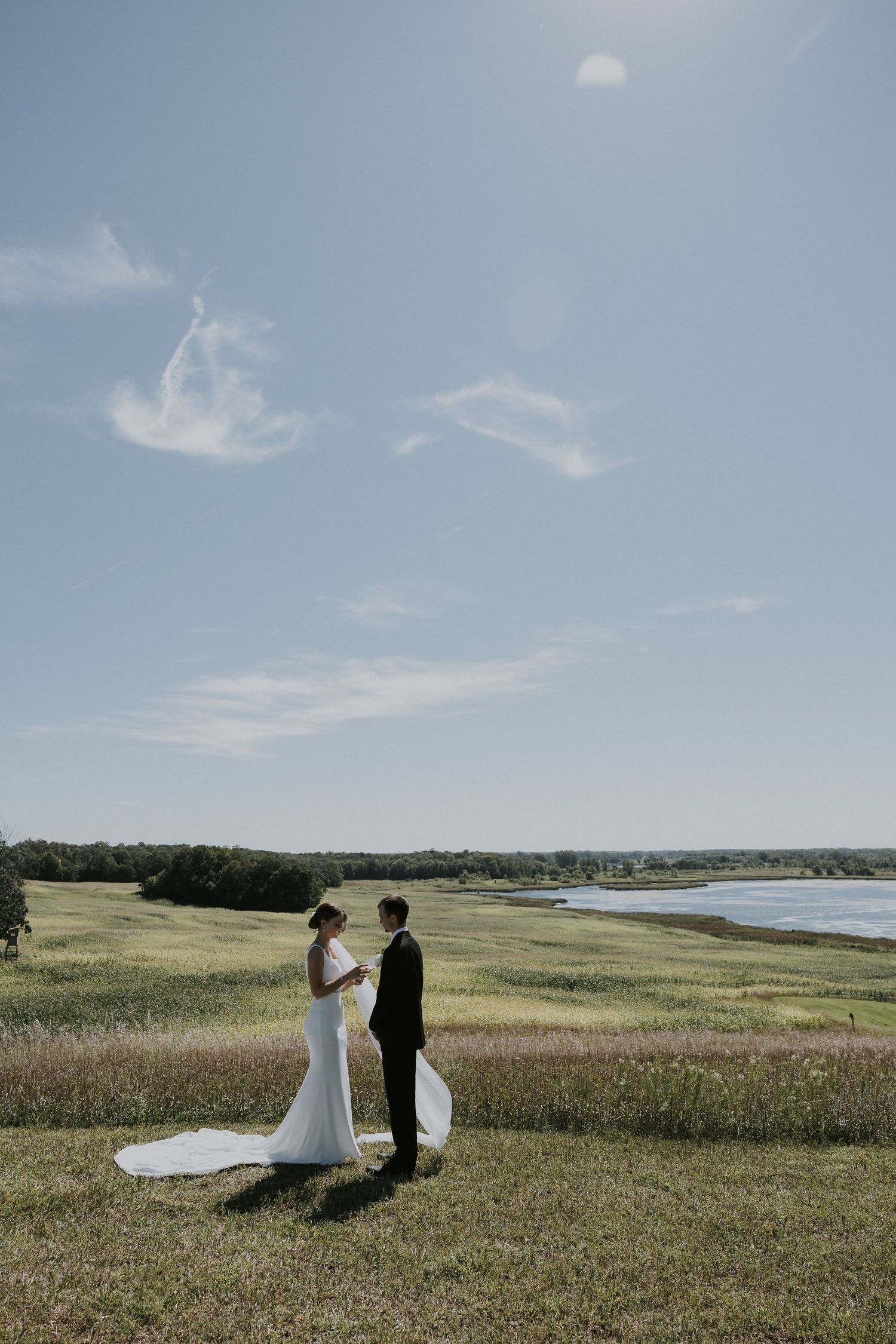 Couple stands together on grassy hilltop overlooking coastal waterfront on sunny day with wispy clouds in blue sky.