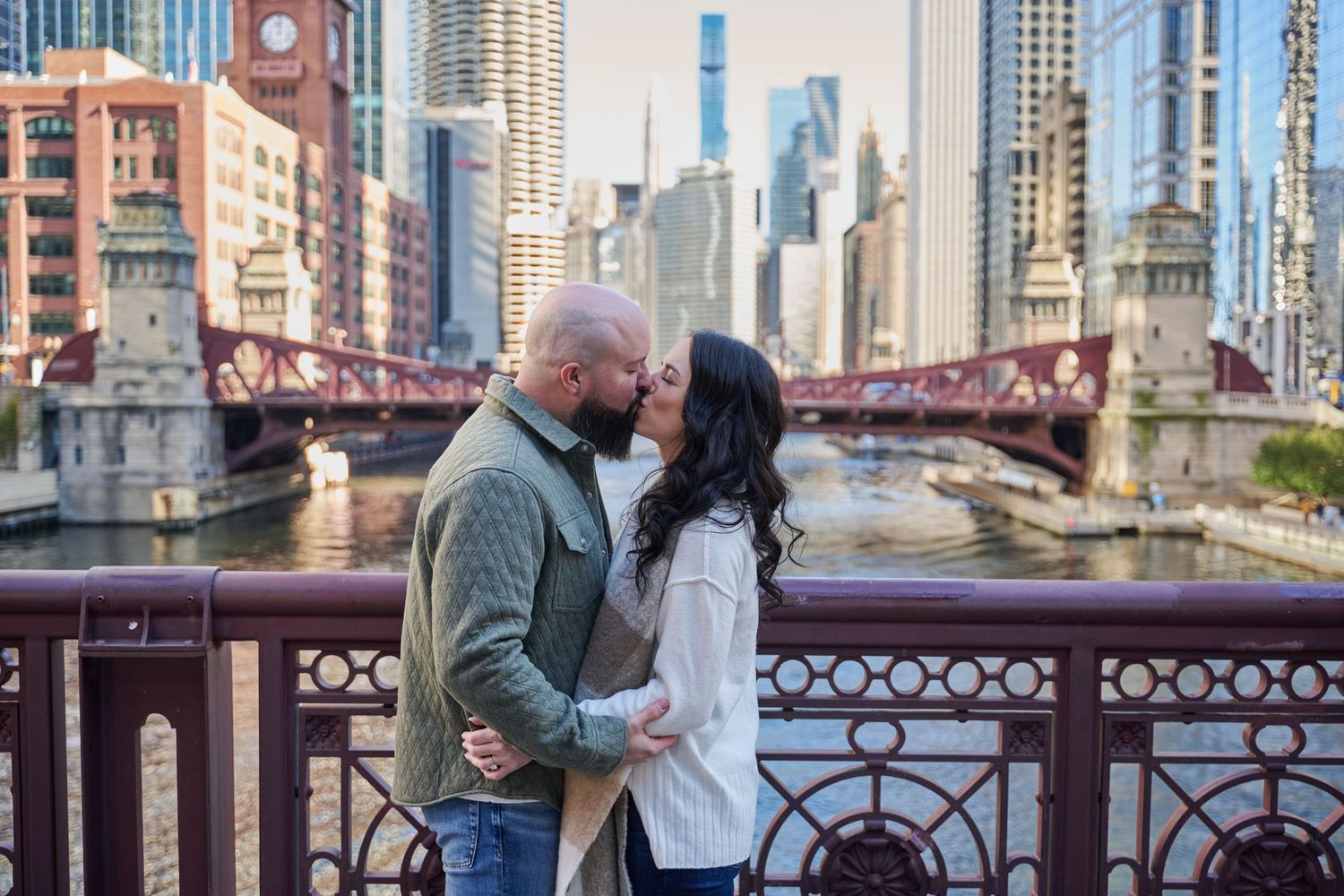 A romantic couple embraces on a bridge over the Chicago River with the city skyline and iconic architecture in the background.