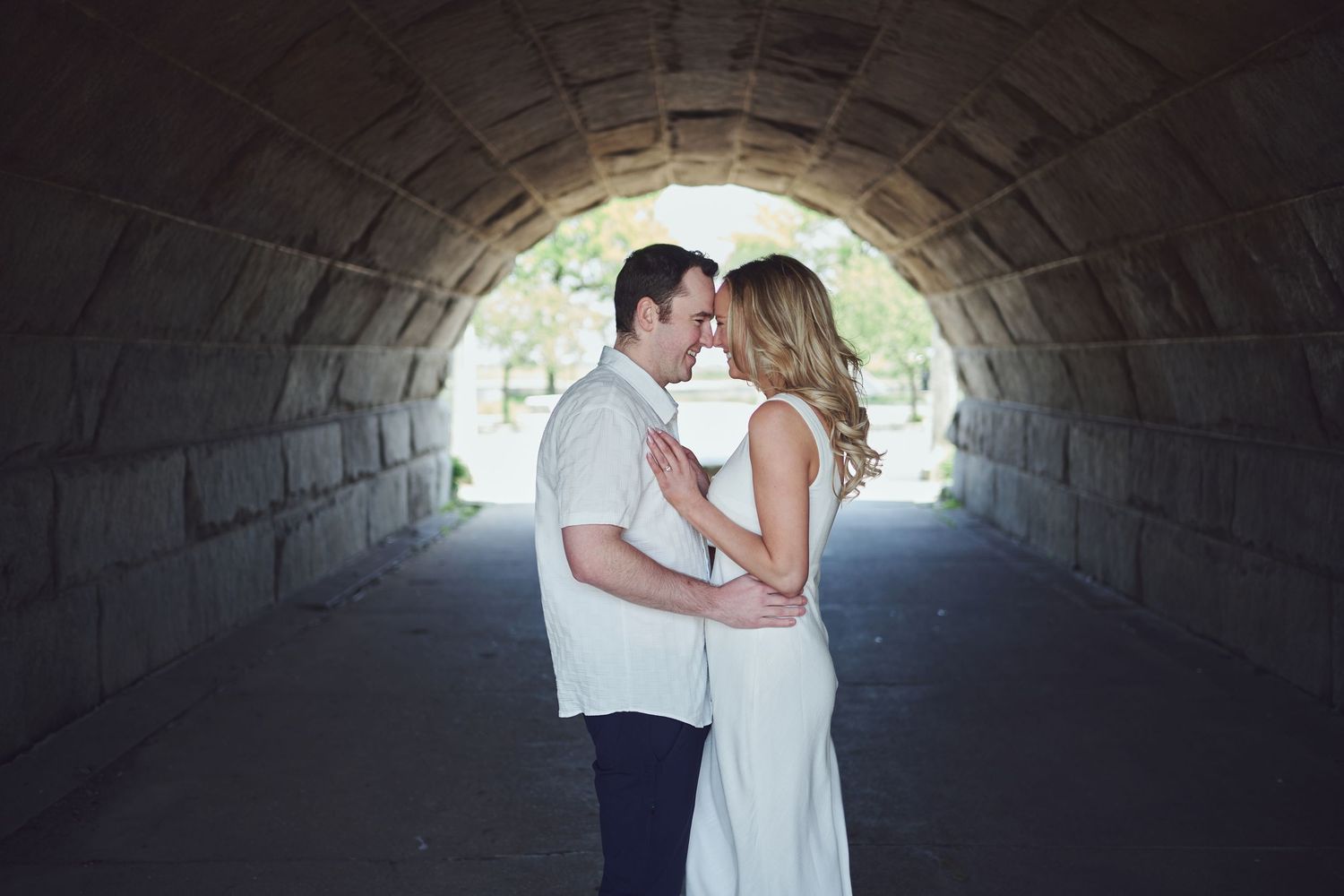 A romantic couple shares a tender kiss under a stone tunnel archway with soft lighting.