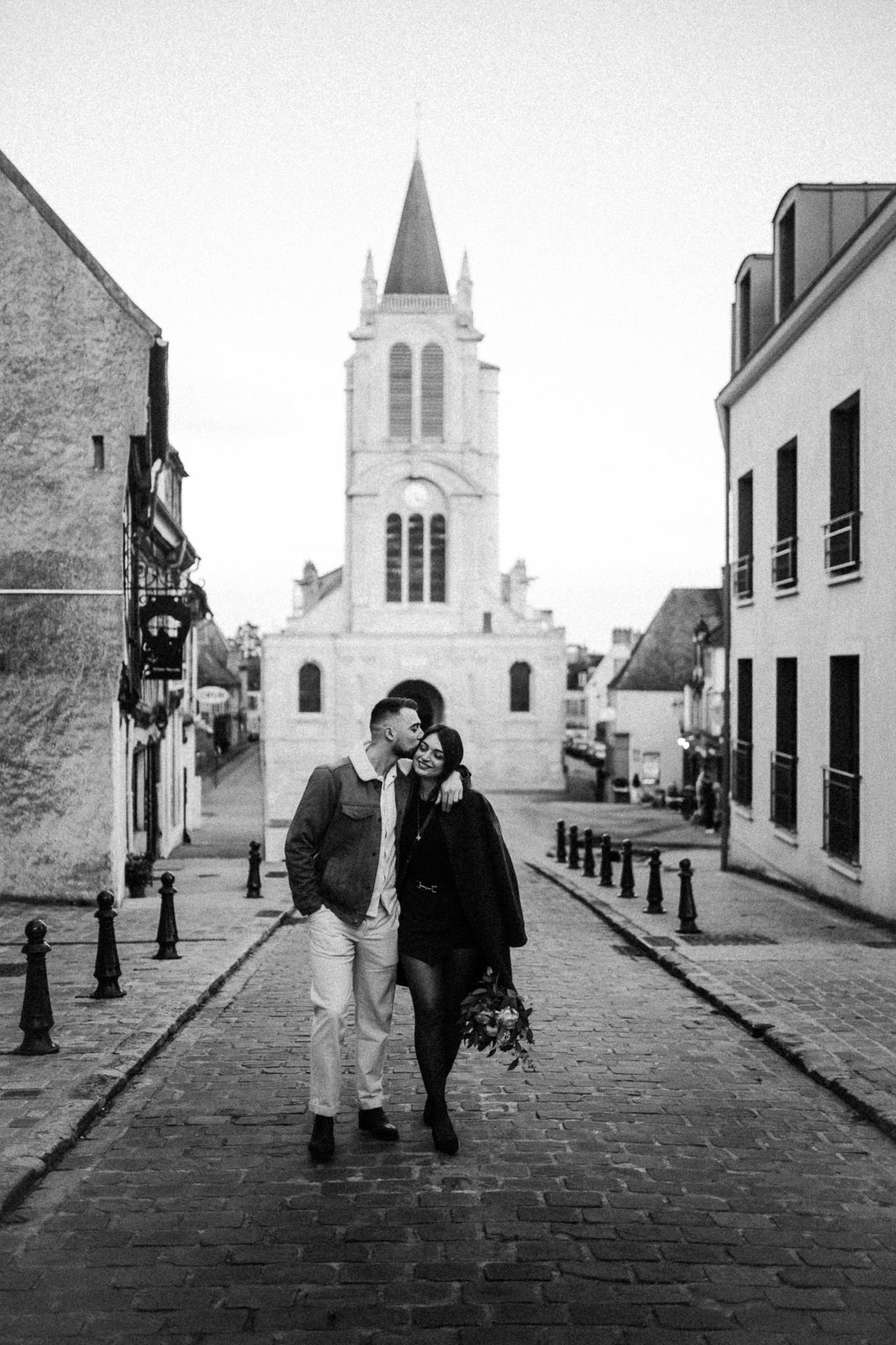 couple marchant dans une rue pavée devant une église à Montfort-l’Amaury lors d’une séance photo
