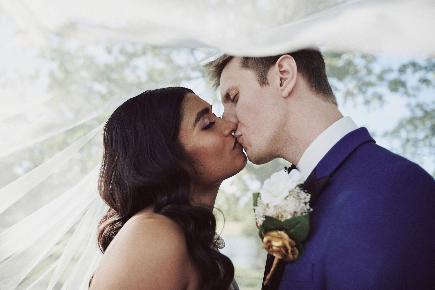 A romantic wedding couple shares a tender kiss under a flowing white veil during their outdoor ceremony.