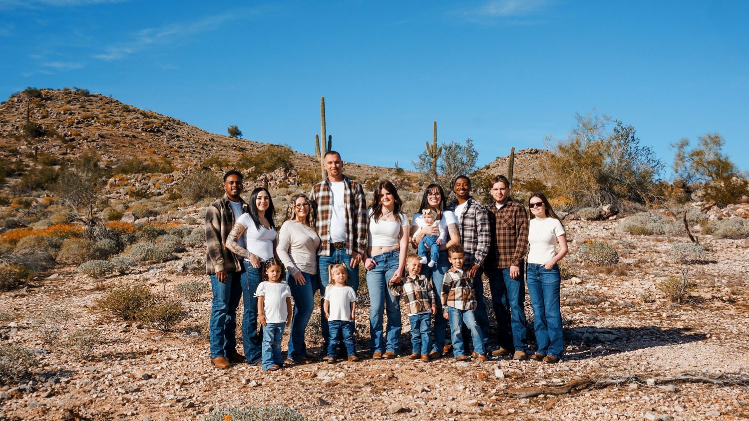 A large group poses together in casual western attire against a desert landscape with rocky hills and blue sky.