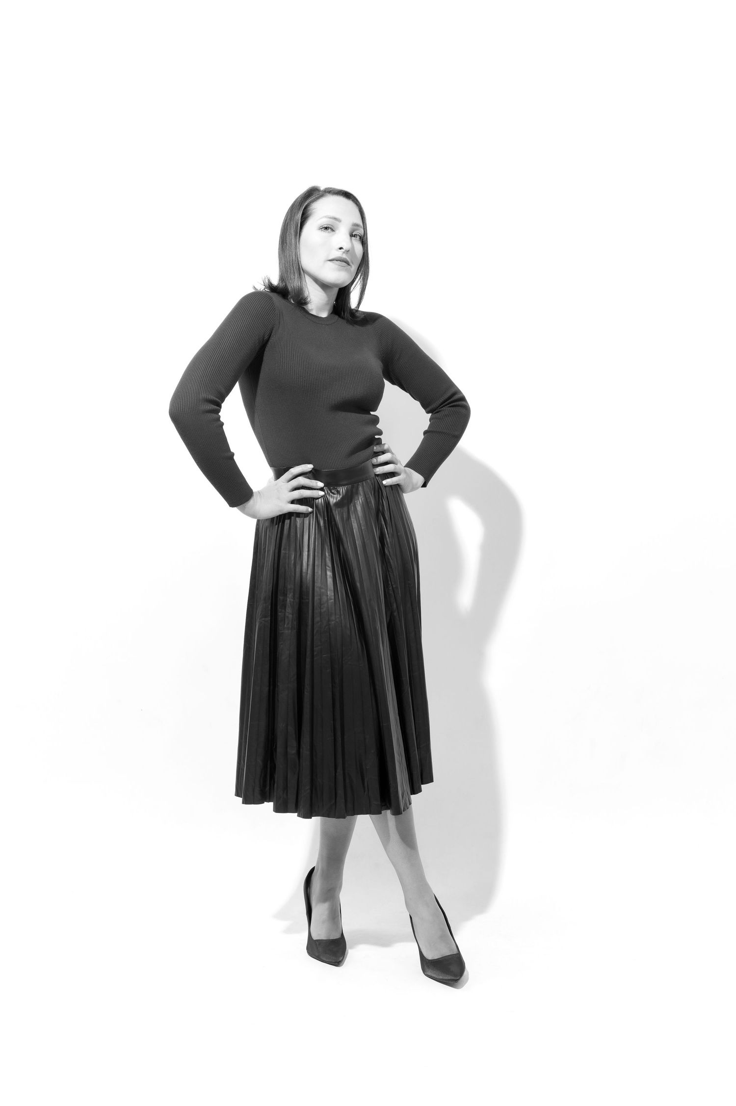 Black and white fashion photo of a figure in a pleated midi skirt and long sleeve top striking a confident pose.