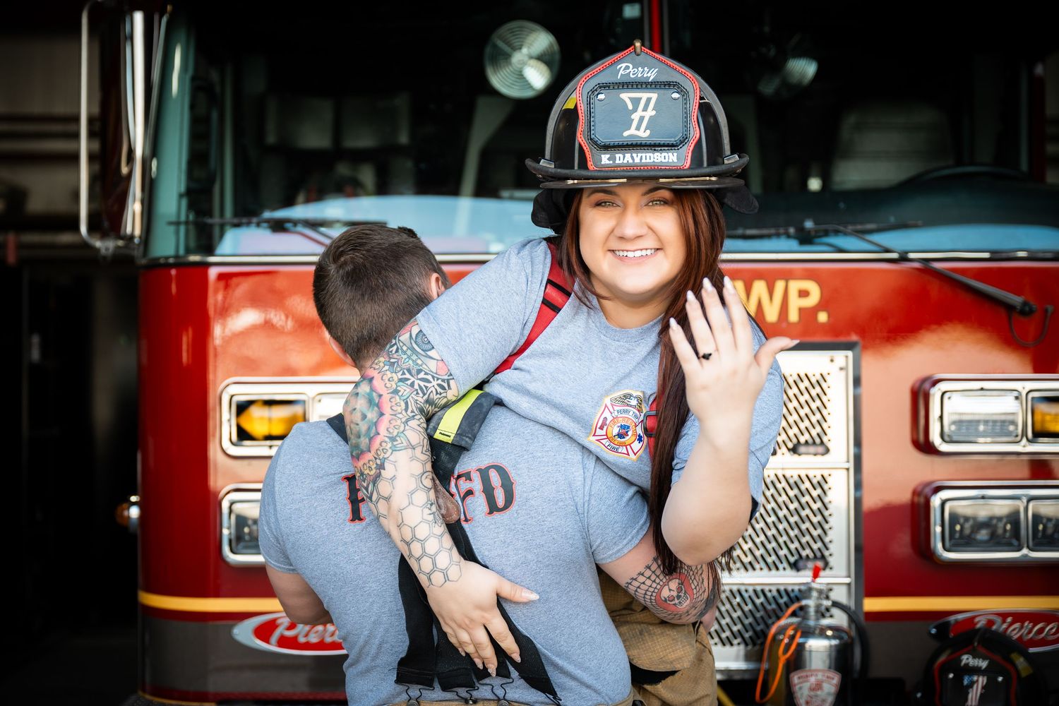 A smiling firefighter in helmet and gear poses in front of a red fire truck at the station.