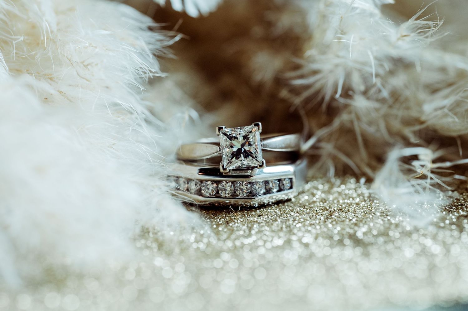 Close-up of a princess-cut diamond engagement ring with wedding band on glittery surface with white feathers.