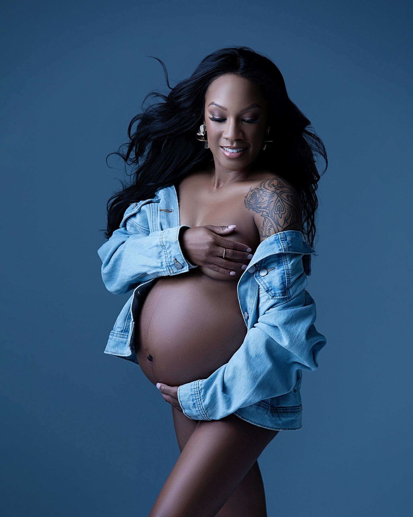 A pregnant woman with long dark hair wearing a denim shirt poses confidently against a blue background.