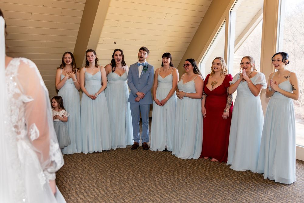 Bridal party in light blue gowns and one red dress reacting emotionally to bride's first look indoors.
