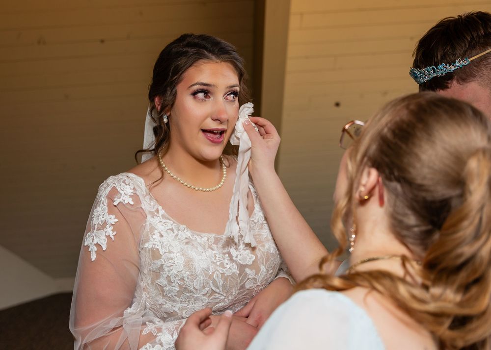 Bride in lace wedding dress gets tears wiped by bridesmaid during emotional wedding day preparation.