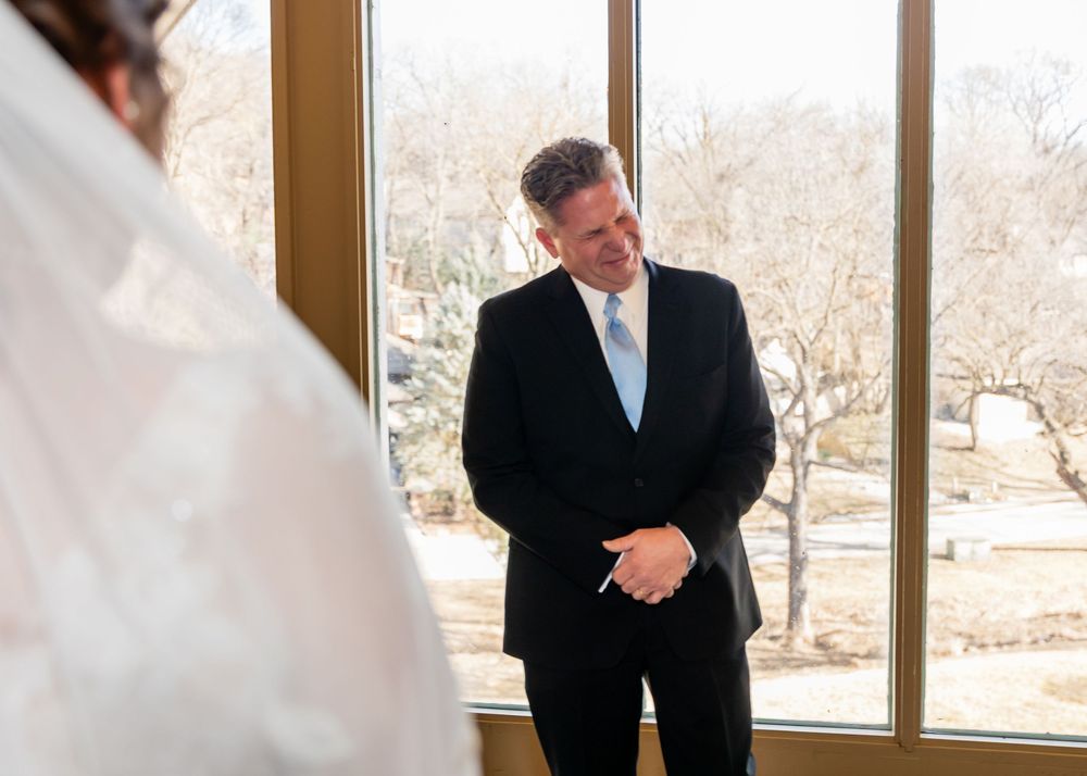 Groom smiling emotionally during first look at bride in elegant wedding venue with large windows.