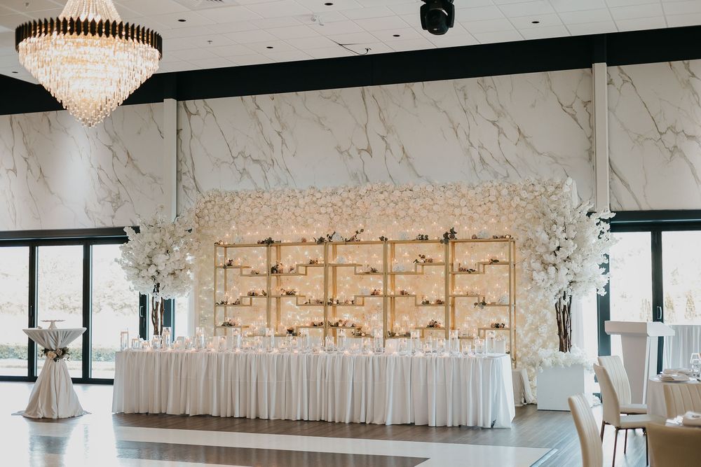Elegant wedding reception hall with white floral backdrop, gold shelving, chandelier, and draped head table.