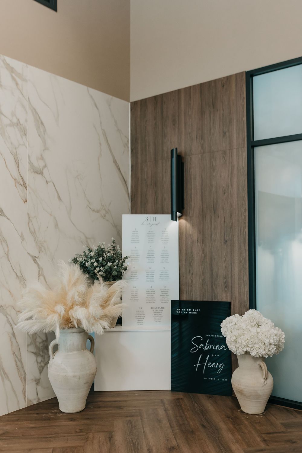 Elegant wedding seating chart display with white ceramic vases, pampas grass, and hydrangeas against marble wall.