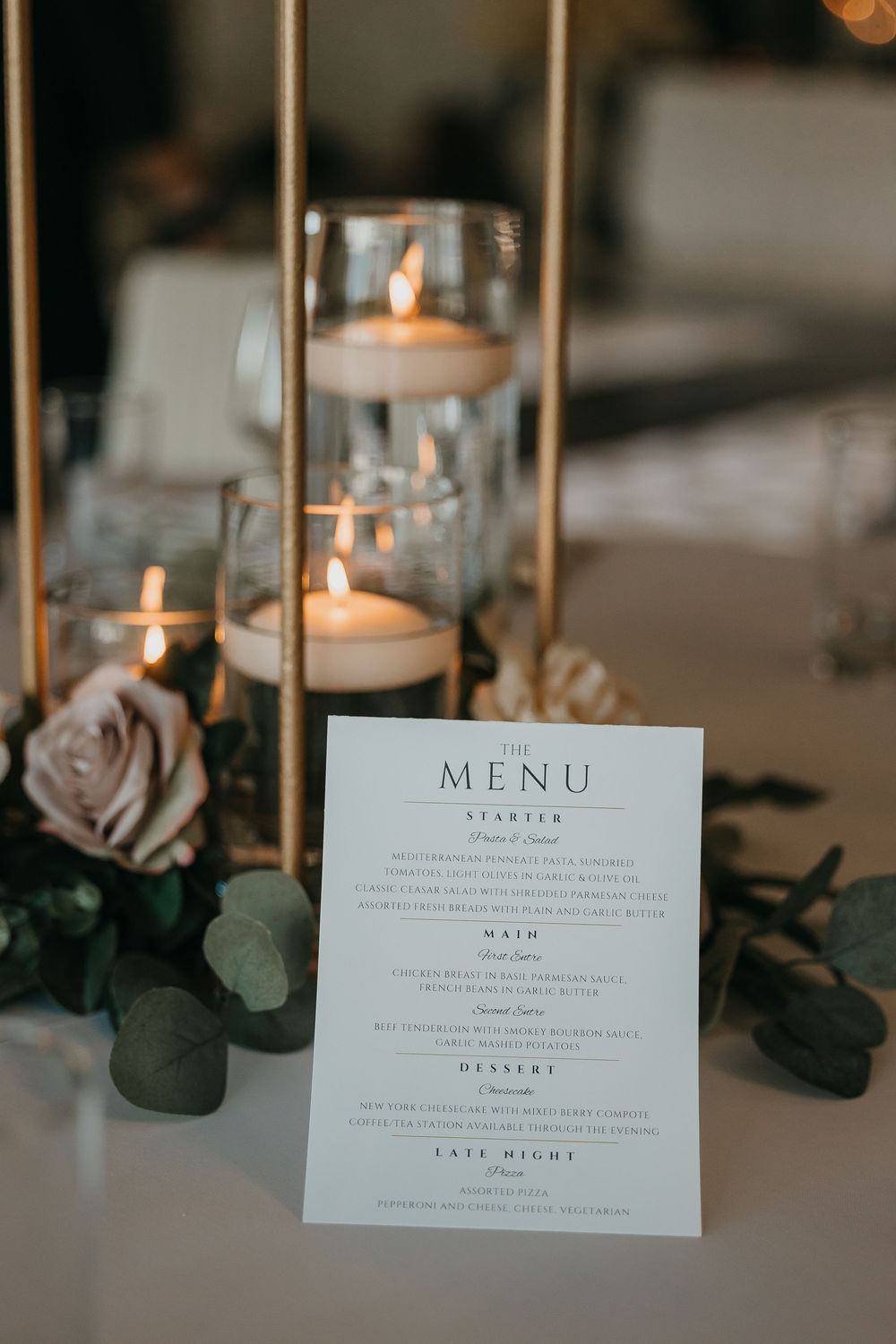 Elegant wedding reception menu card displayed beside gold candle holders with floating candles and blush roses.