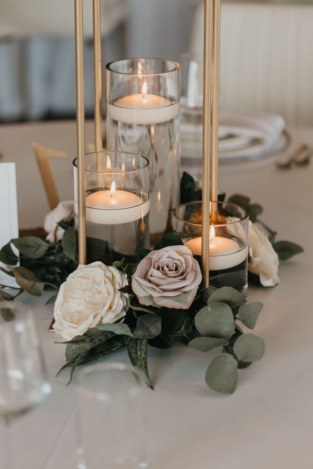 Elegant wedding centerpiece with floating candles in glass cylinders, gold rods, and white roses with eucalyptus greenery.
