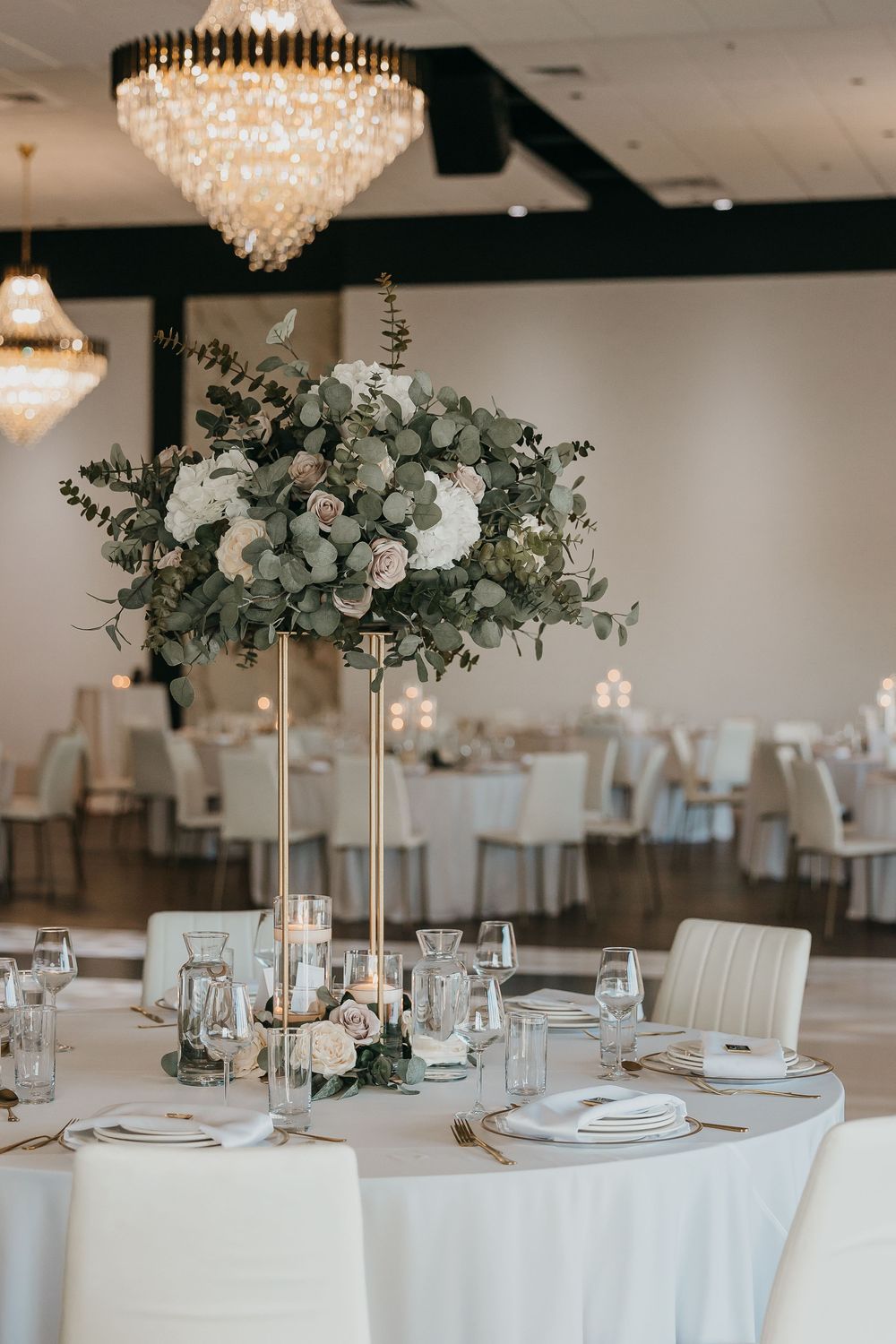 Elegant wedding reception table with tall gold floral centerpiece, white roses, eucalyptus, crystal chandeliers, and white draped chairs.