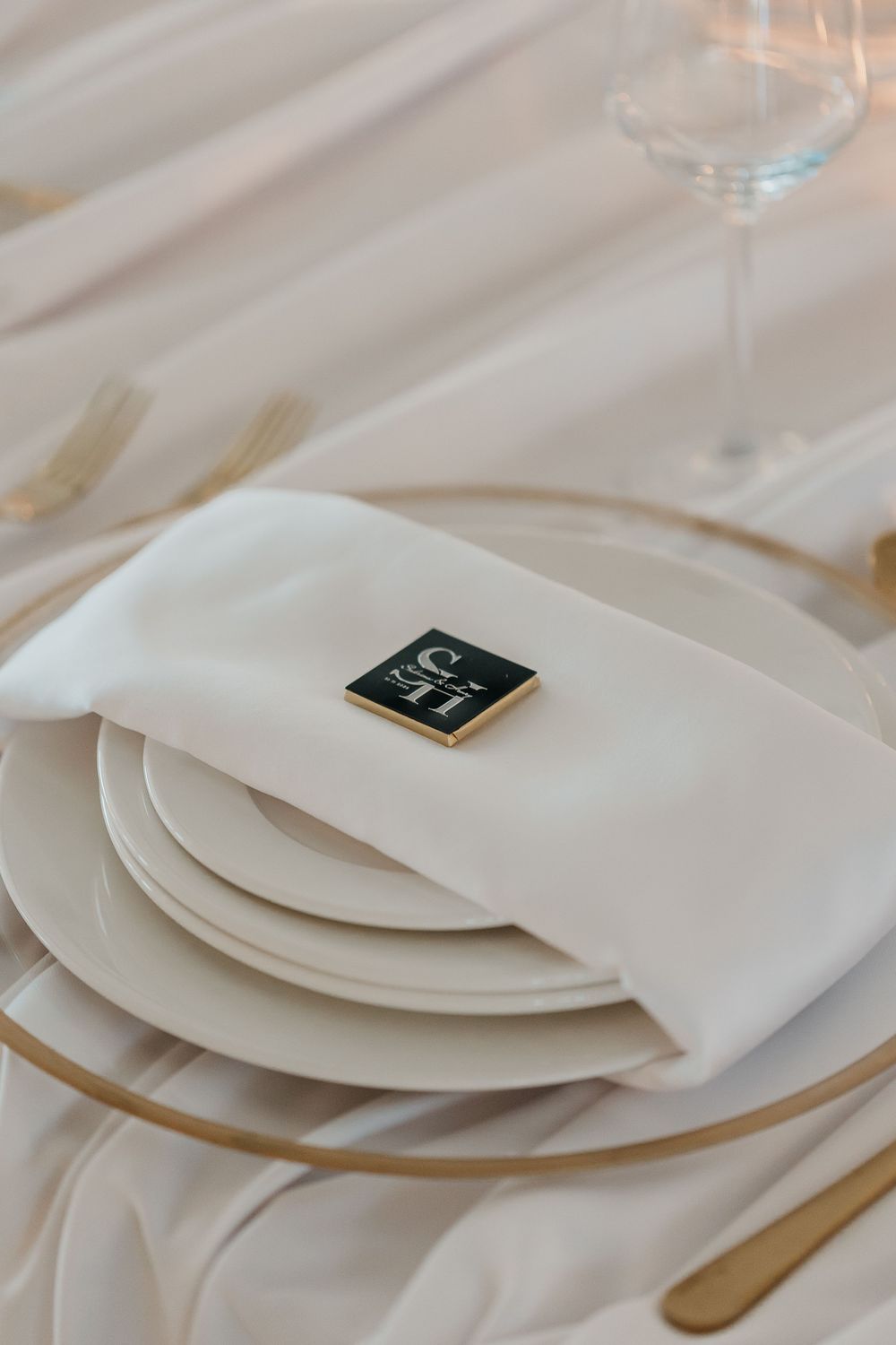 White wedding place setting with gold cutlery, monogram card on folded napkin, and crystal wine glass.