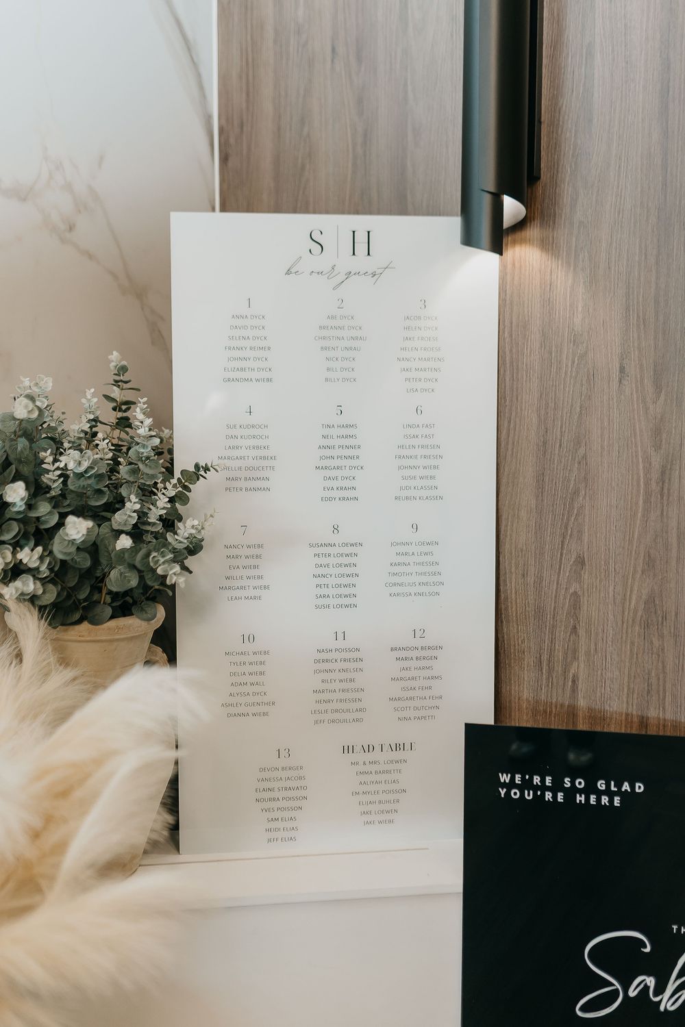 Elegant wedding seating chart sign with monogram, displayed beside greenery and a fluffy white decoration.