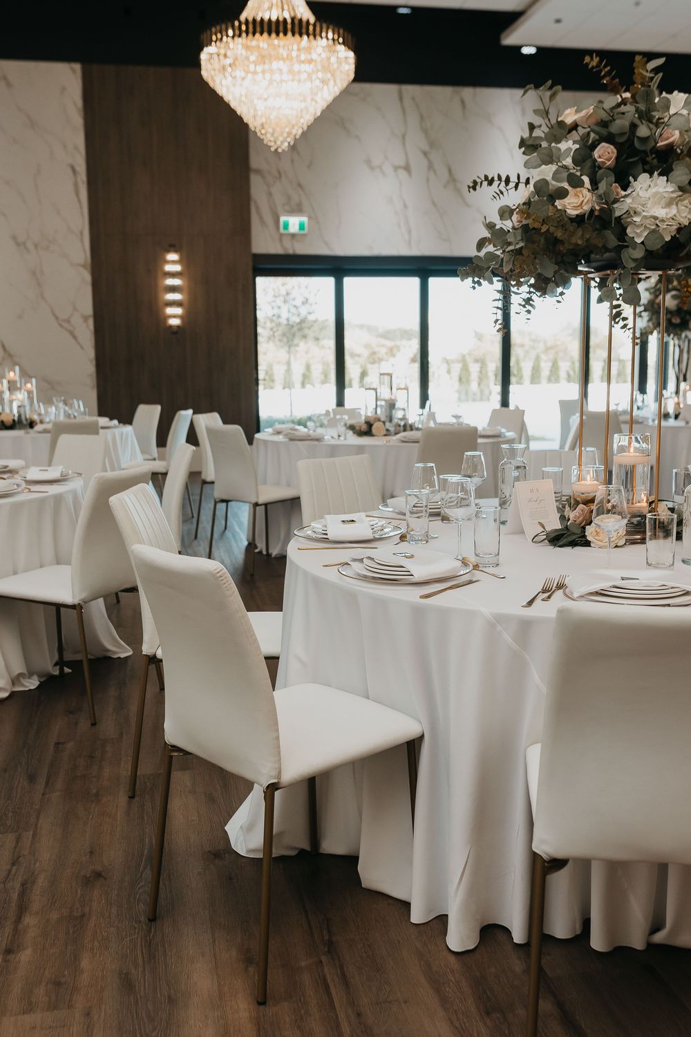 Elegant wedding reception hall with white round tables, ghost chairs, crystal chandelier, and tall floral centerpieces.