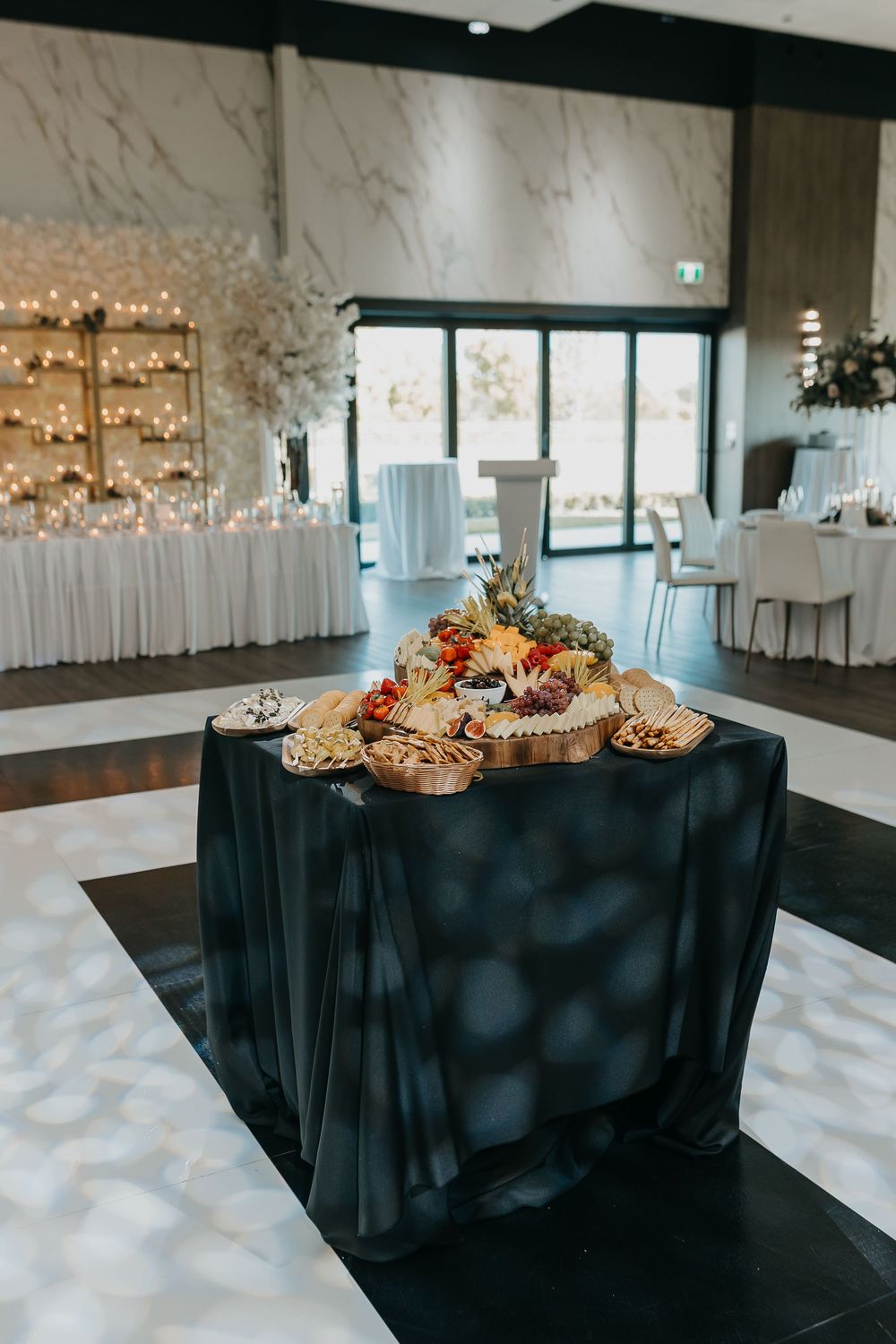 Elegant wedding reception grazing table with dark linen, charcuterie display, and candlelit floral backdrop on black and white dance floor.