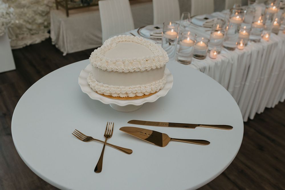 White wedding cake on round table with gold fork, knife, and cake server, candles glowing in background.
