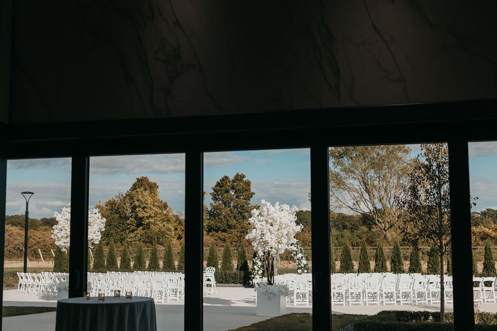 Indoor view through large windows of an elegant outdoor wedding ceremony setup with white chairs and floral arrangements.