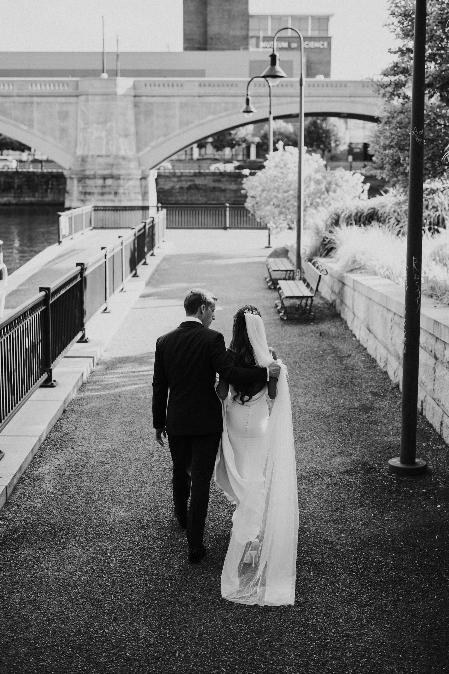 Black and white photo of couple walking along riverside path near historic architecture.