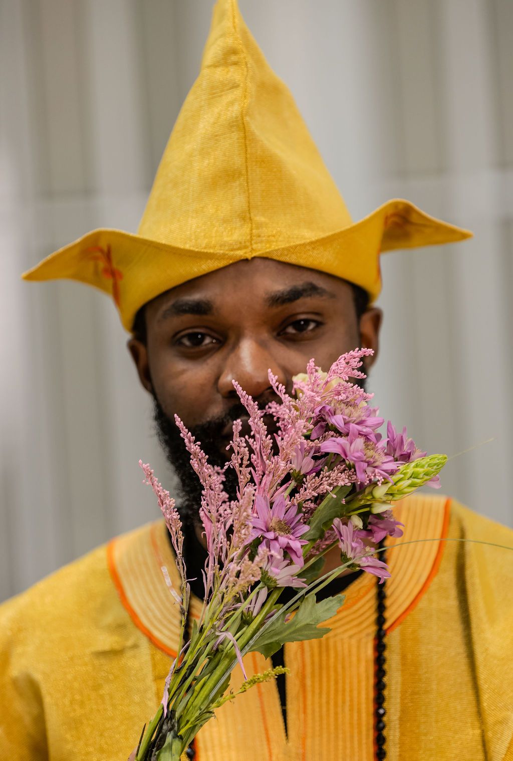 Man in vibrant yellow African attire and pointed hat holding pink wildflowers near his face.