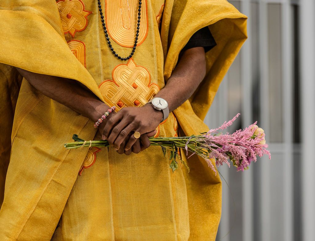 Close-up of hands holding pink wildflowers wearing a watch and bracelet in yellow embroidered African kaftan.