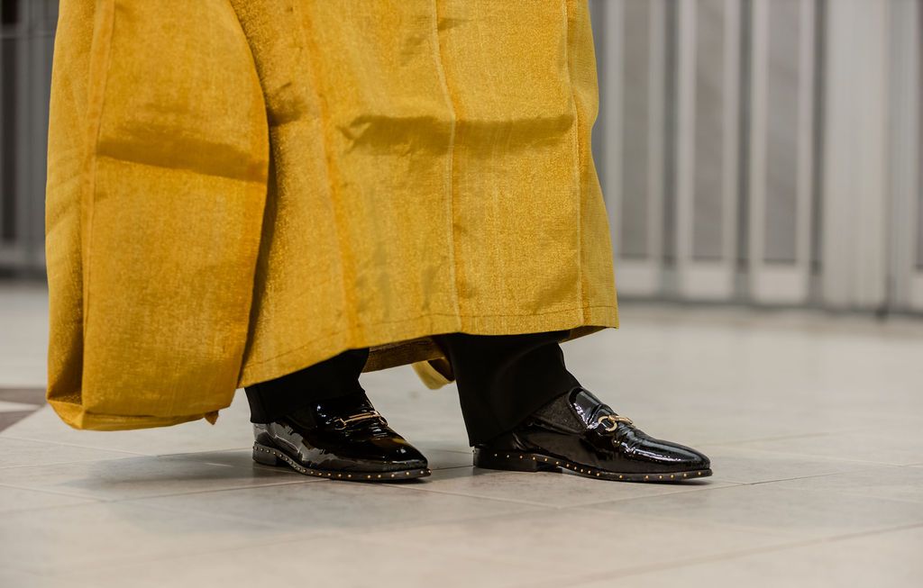 Black patent leather loafers with gold hardware worn with black trousers under a yellow traditional African agbada robe.