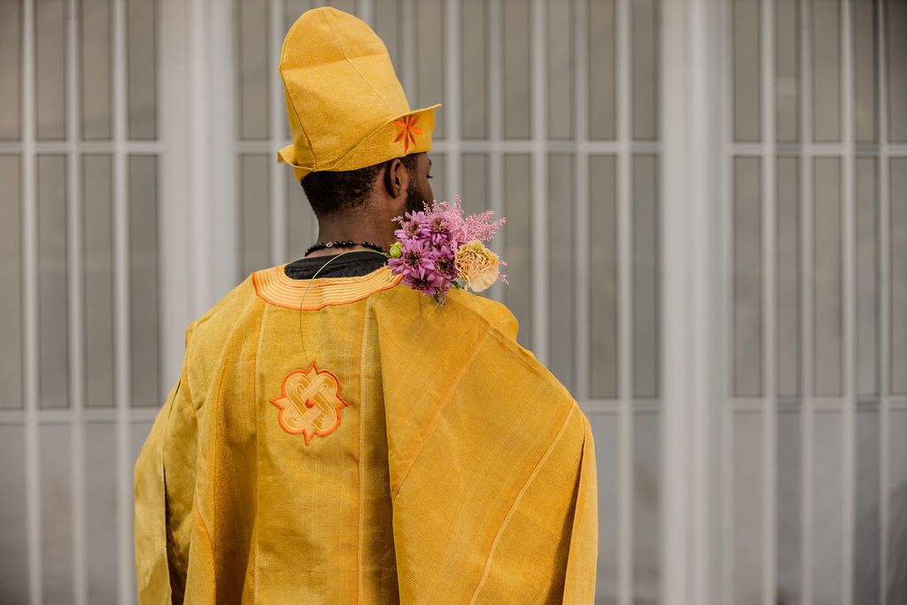 Back view of Black man in yellow embroidered African agbada with tall hat decorated with purple flowers.