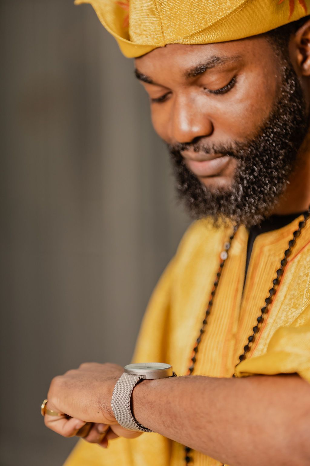 Bearded Black man in yellow African attire checking silver wristwatch while looking down thoughtfully.