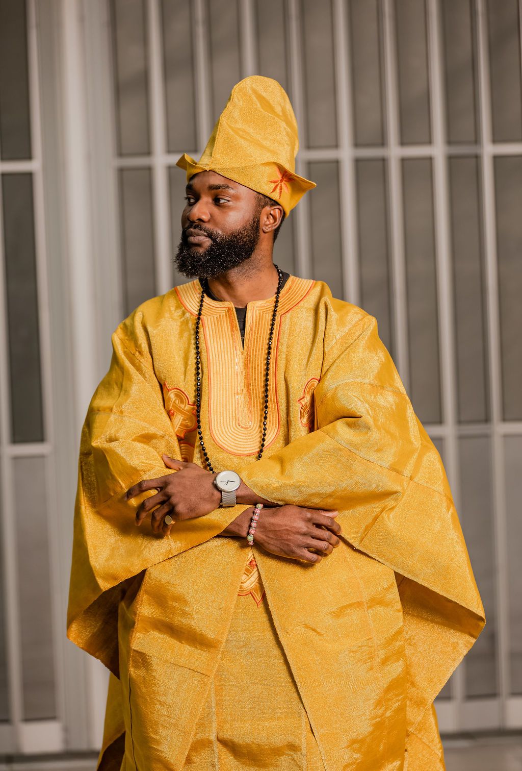Stylish Black man in full yellow traditional Nigerian agbada outfit with tall fila hat and silver watch posing confidently.