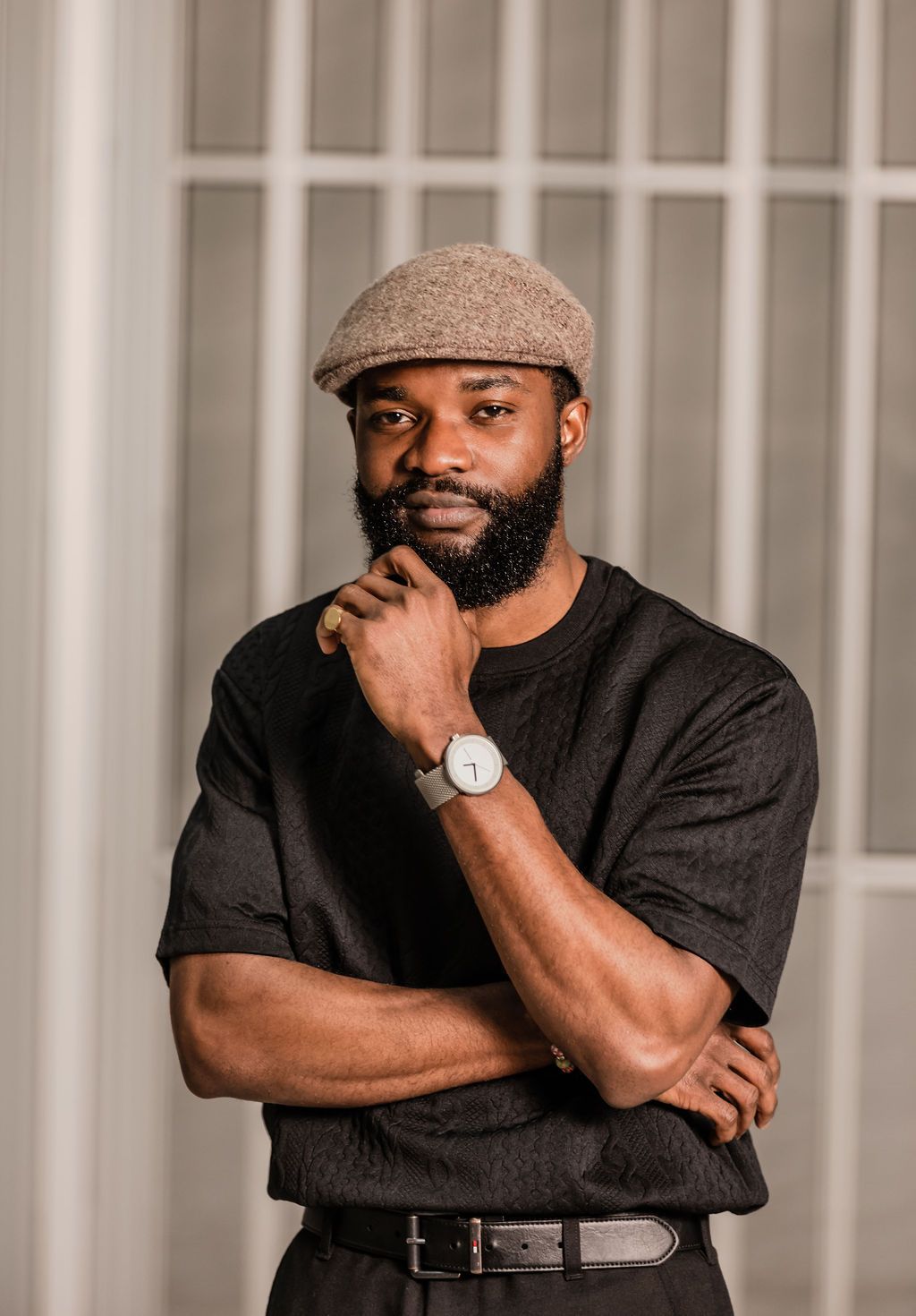 Confident bearded Black man in black outfit wearing grey flat cap and silver watch with arms crossed.