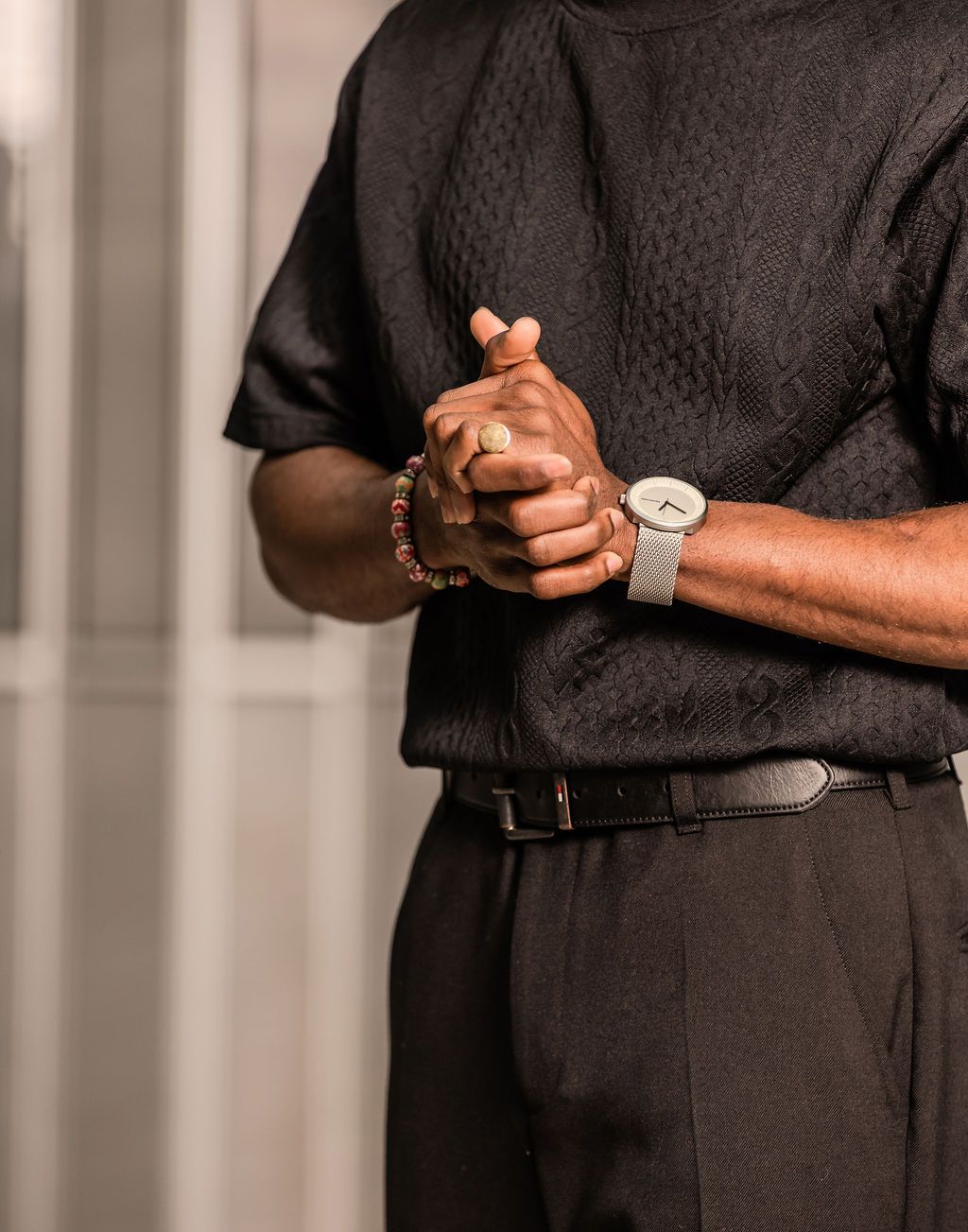 Close-up of Black man in black textured shirt clasping hands showing beaded bracelet and minimalist silver watch.