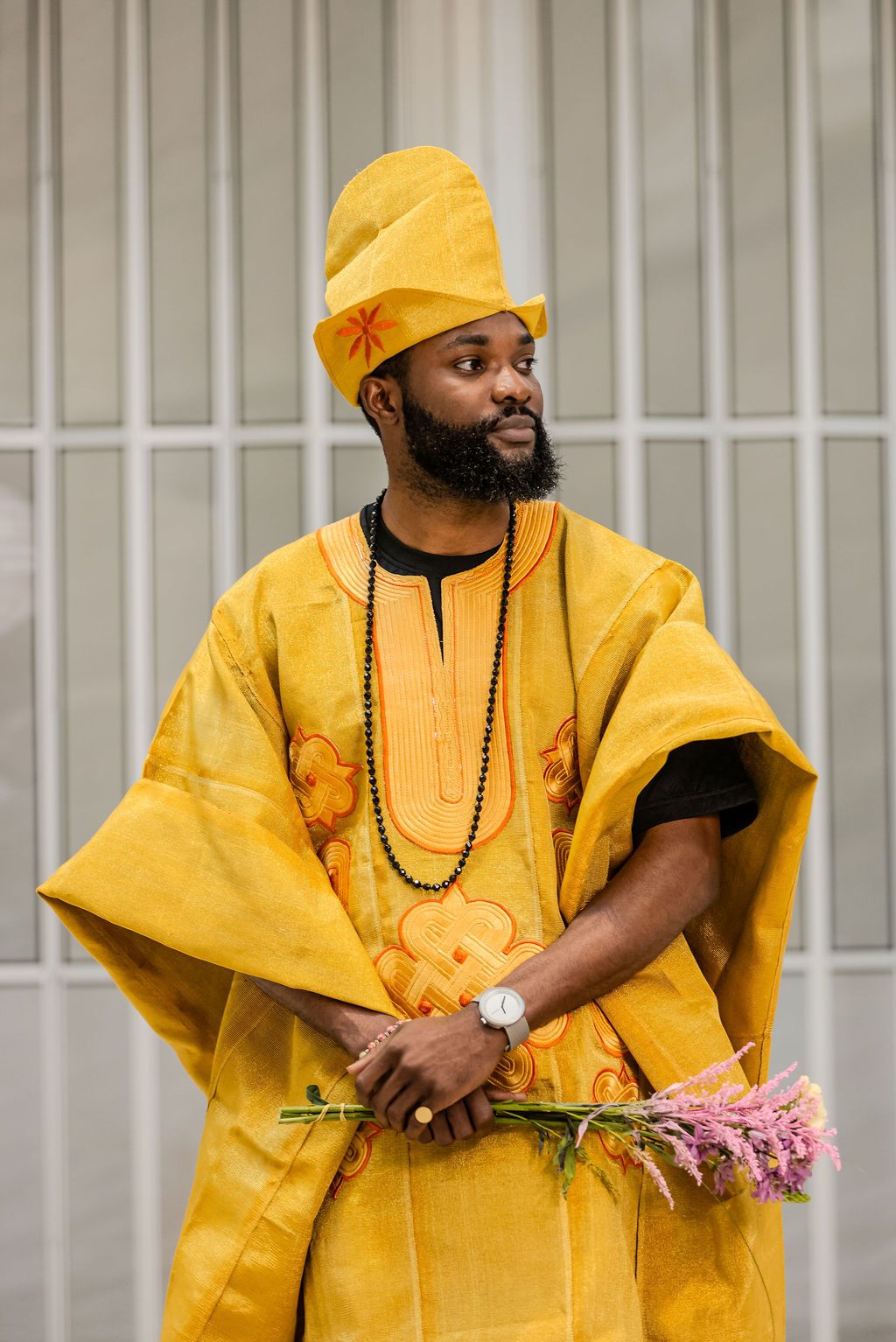 Bearded man in traditional yellow African agbada robe and gele hat holding pink flowers outdoors.
