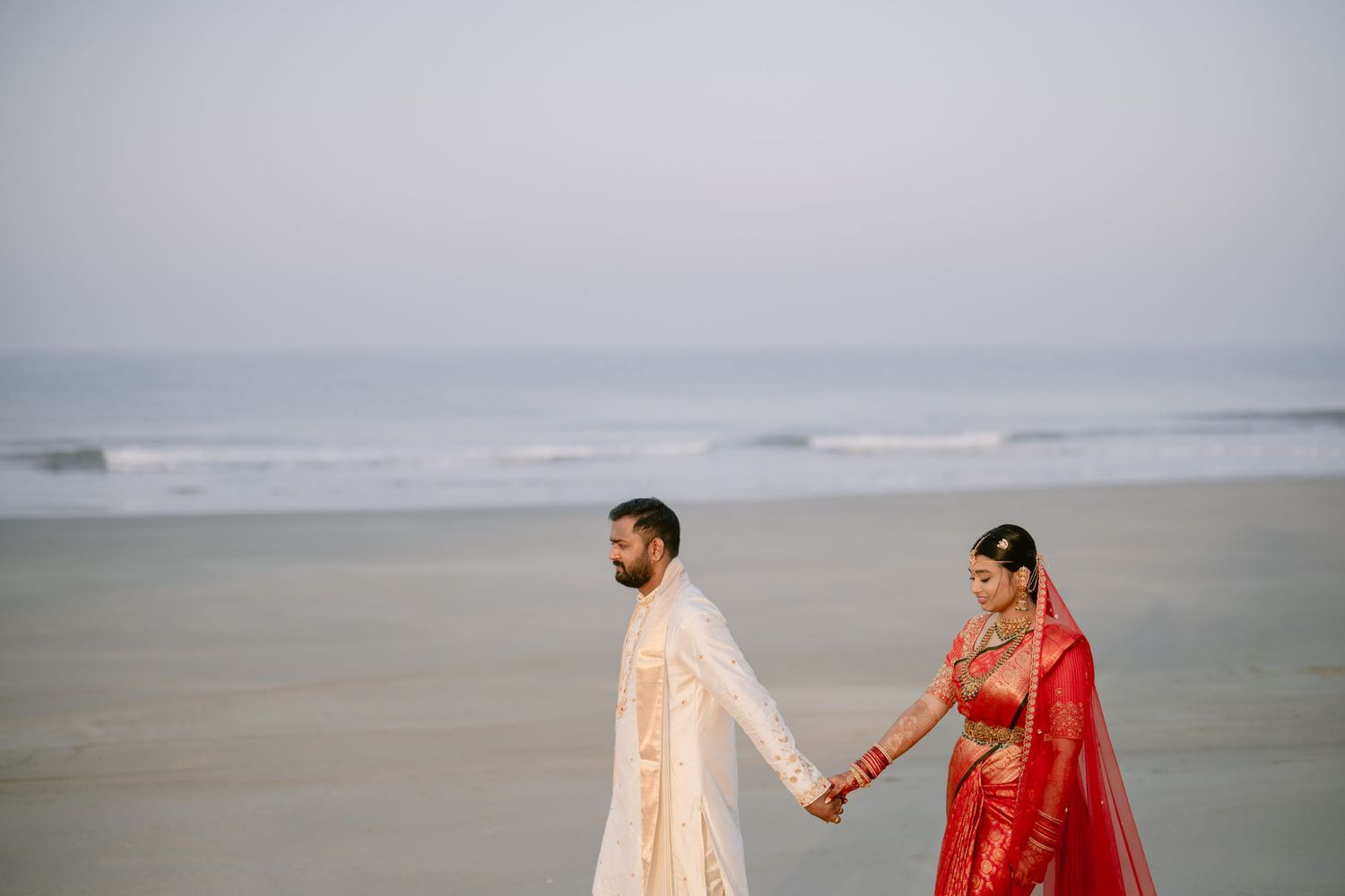 Indian bride in red saree and groom in cream sherwani hold hands walking along a serene beach at dawn.