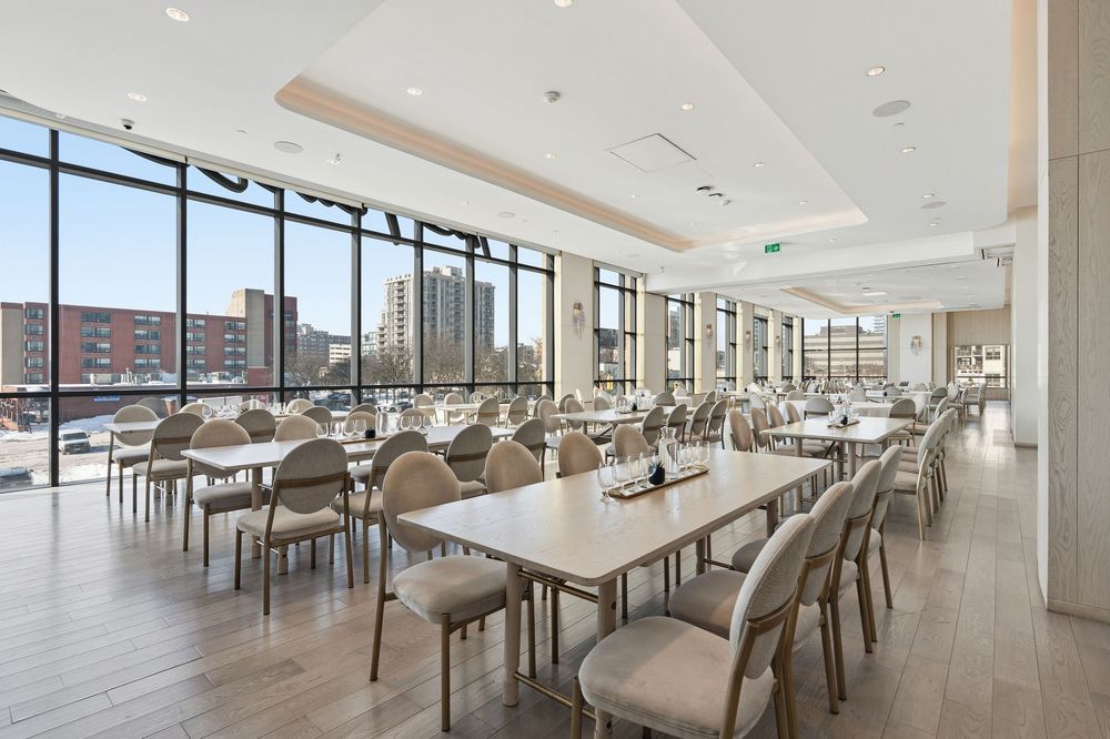 Modern office cafeteria with long white tables, chairs and floor-to-ceiling windows offering panoramic city views.