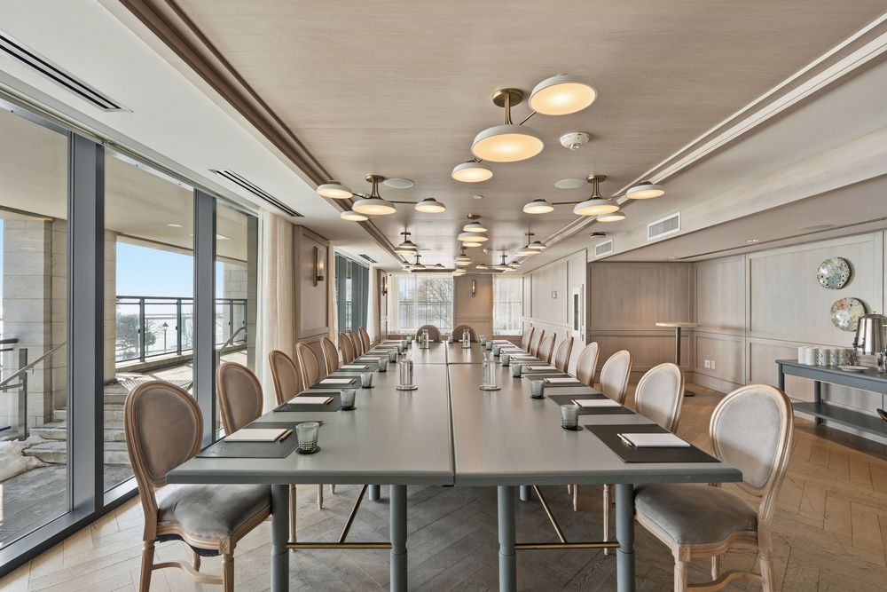 Executive boardroom with long conference table, upholstered chairs, and modern lighting fixtures along a window-lined wall.