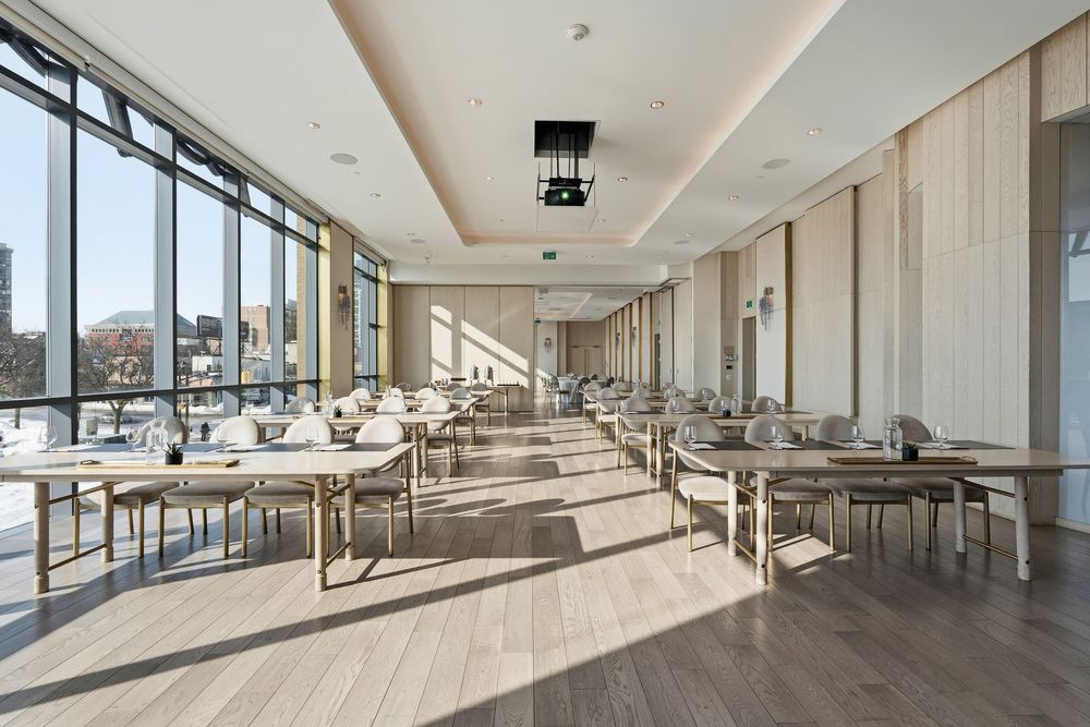 Modern conference room with floor-to-ceiling windows, wooden floors, and multiple tables arranged in rows with natural lighting.