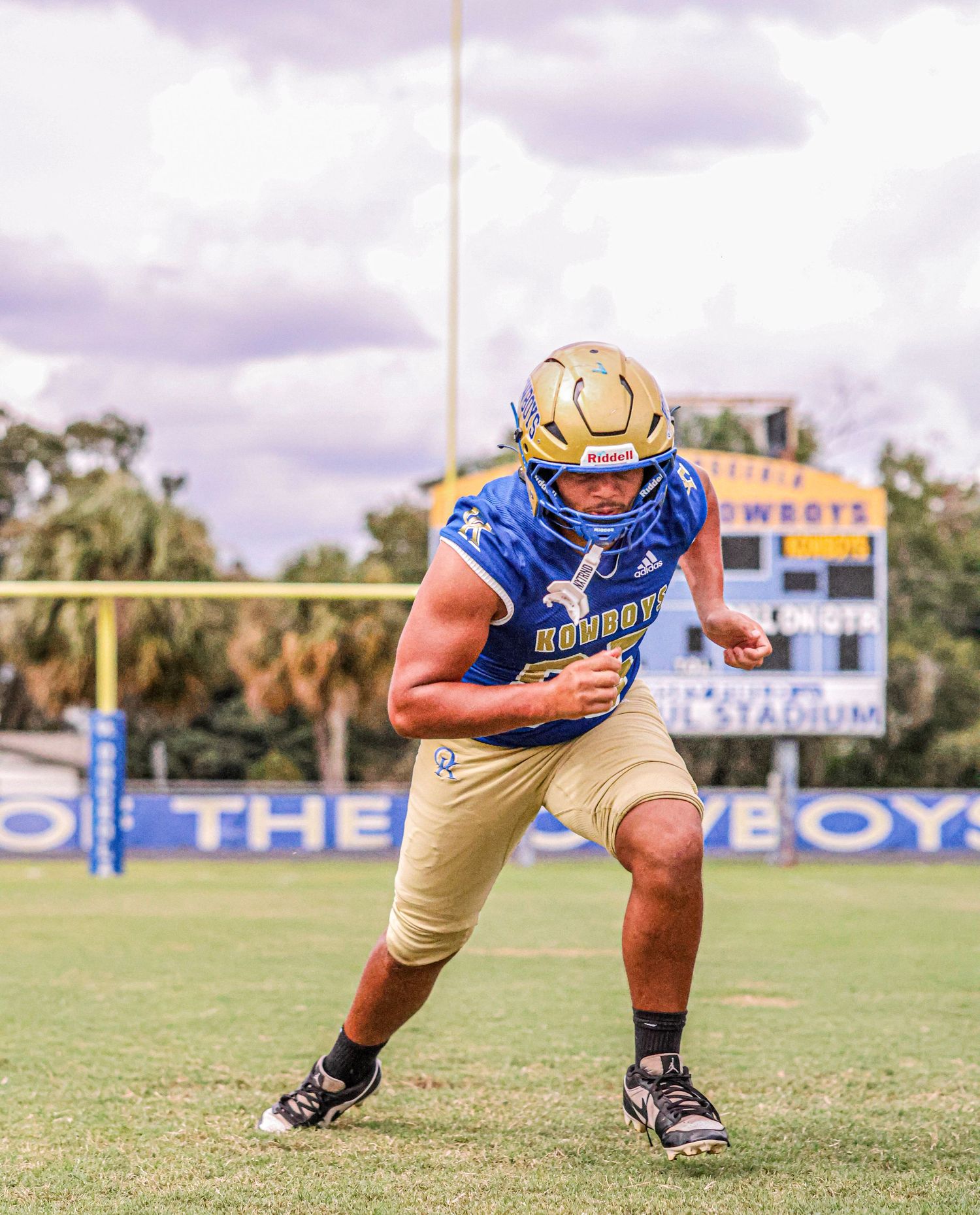 A football player in blue and gold uniform crouches low in a defensive stance on the playing field.