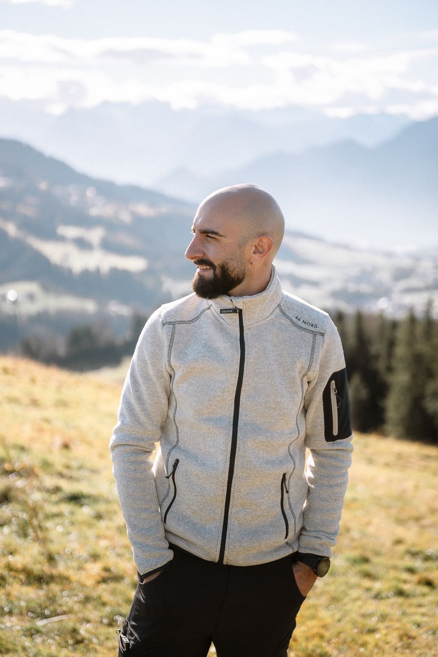 Ein lächelnder, bärtiger Mann in heller Fleecejacke steht auf einer Wiese und blickt über eine sonnige Schweizer Berglandschaft.