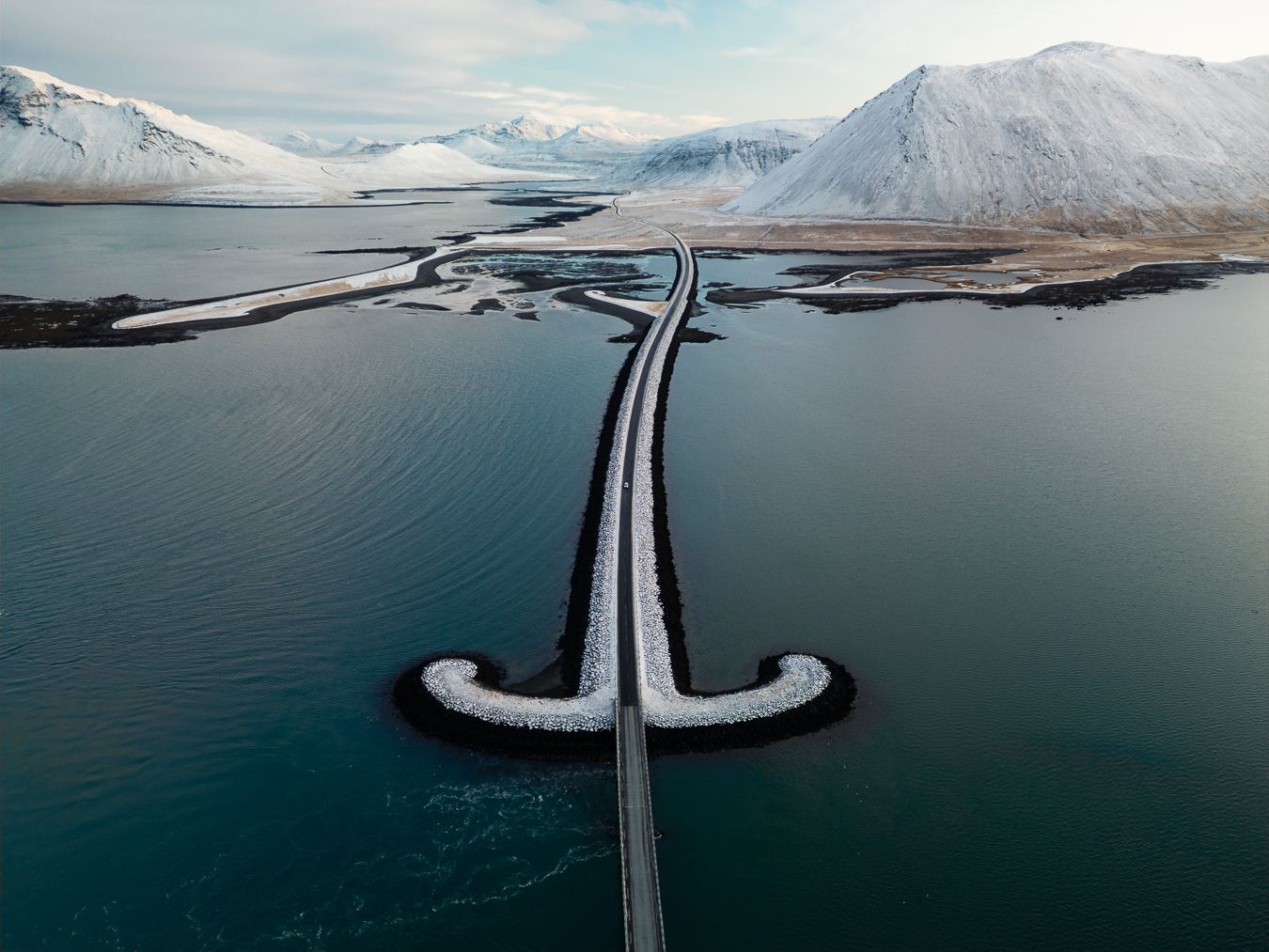 Aerial view of snow-covered bridge curving through icy blue fjord with mountains.